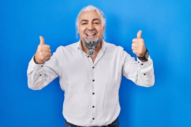 Middle age man with grey hair standing over blue background success sign doing positive gesture with hand, thumbs up smiling and happy. cheerful expression and winner gesture. 
