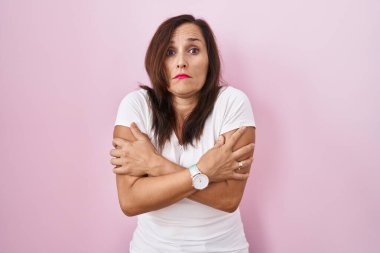 Middle age brunette woman standing over pink background shaking and freezing for winter cold with sad and shock expression on face 