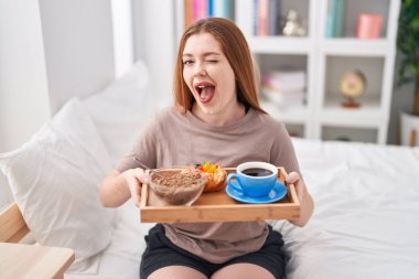Redhead woman wearing pajama holding breakfast tray winking looking at the camera with sexy expression, cheerful and happy face. 