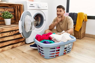 Young handsome man putting dirty laundry into washing machine looking confident at the camera with smile with crossed arms and hand raised on chin. thinking positive. 