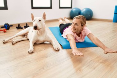 Genç beyaz kadın gülümsüyor spor merkezinde köpekle esniyor.
