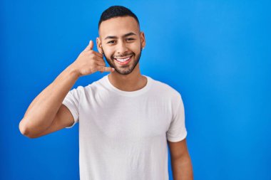 Young hispanic man standing over blue background smiling doing phone gesture with hand and fingers like talking on the telephone. communicating concepts. 