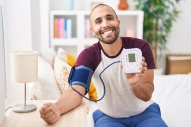 Young latin man measuring pulse using tensiometer at bedroom