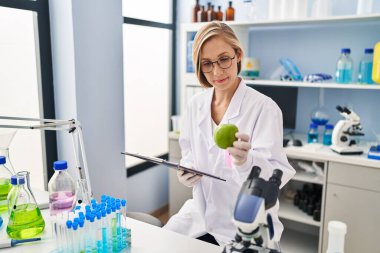 Young blonde woman scientist holding apple reading report at laboratory