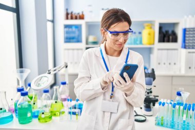 Laboratuvarda akıllı telefon kullanan genç İspanyol kadın bilim adamı üniforması giyiyor.