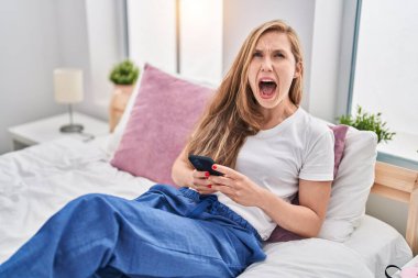 Young blonde woman using smartphone on bed angry and mad screaming frustrated and furious, shouting with anger looking up. 