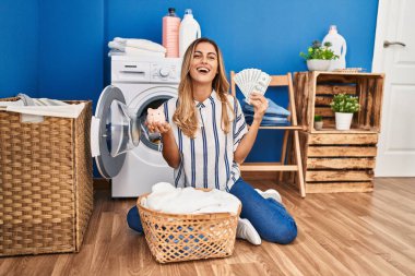 Young blonde woman doing laundry saving money smiling and laughing hard out loud because funny crazy joke. 