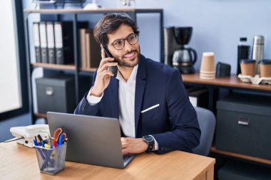 Genç İspanyol iş adamı bilgisayarını kullanıyor ve ofisteki akıllı telefondan konuşuyor.
