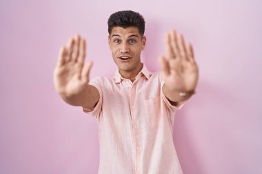 Young hispanic man standing over pink background doing stop gesture with hands palms, angry and frustration expression 