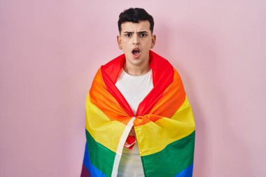 Non binary person holding rainbow lgbtq flag scared and amazed with open mouth for surprise, disbelief face 