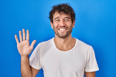 Mavi arka planda dikilen İspanyol genç adam beş numarayı gösterip işaret ederken kendinden emin ve mutlu gülümsüyor.. 