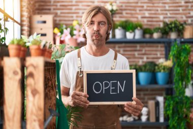 Caucasian man with mustache working at florist holding open sign puffing cheeks with funny face. mouth inflated with air, catching air. 