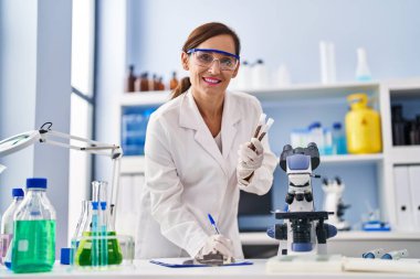 Middle age woman wearing scientist uniform writing on clipboard holding test tubes at laboratory