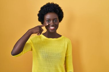 African young woman standing over yellow studio pointing with hand finger to face and nose, smiling cheerful. beauty concept 