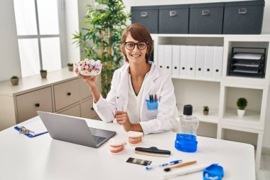 Brunette dentist woman holding sweets looking positive and happy standing and smiling with a confident smile showing teeth 