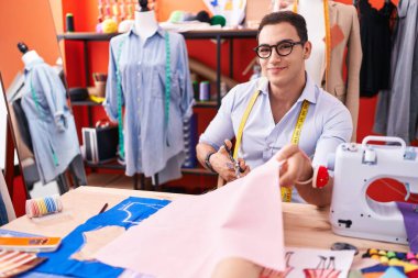 Young hispanic man tailor smiling confident cutting cloth at atelier