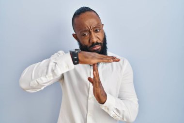 African american man standing over blue background doing time out gesture with hands, frustrated and serious face 
