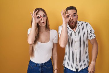 Young couple standing over yellow background doing ok gesture shocked with surprised face, eye looking through fingers. unbelieving expression. 