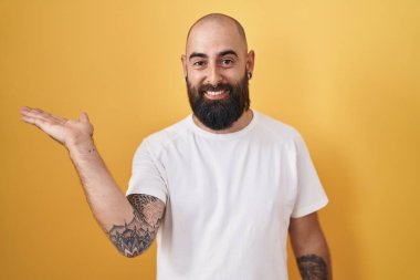 Young hispanic man with beard and tattoos standing over yellow background smiling cheerful presenting and pointing with palm of hand looking at the camera. 