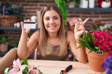 Young blonde woman working at florist shop holding credit card smiling happy pointing with hand and finger to the side 