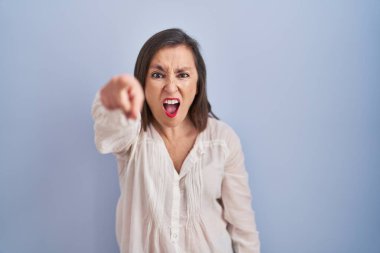 Orta yaşlı Latin bir kadın mavi arka planda dikiliyor. Kameraya kızgın ve sinirli olduğunu gösteriyor. 