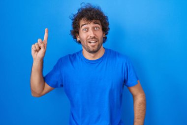Mavi arka planda duran İspanyol genç adam başarılı bir fikirle işaret ediyor. Heyecanlı ve mutlu. Bir numara.. 
