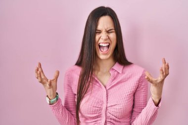 Young hispanic woman standing over pink background celebrating mad and crazy for success with arms raised and closed eyes screaming excited. winner concept 