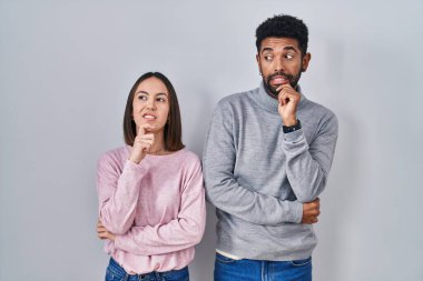 Young hispanic couple standing together thinking worried about a question, concerned and nervous with hand on chin 