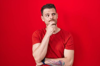 Elleri çenesinde kırmızı arka planda dikilmiş soru soran, düşünceli bir İspanyol genç. Gülümseyen ve düşünceli bir yüz. şüphe kavramı. 