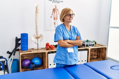 Klinikte fizyoterapist üniforması giyen orta yaşlı sarışın kadın.