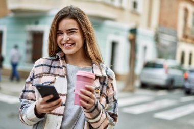 Genç sarışın kadın gülümsüyor. Akıllı telefon kullanıyor. Sokakta kahve tutuyor.