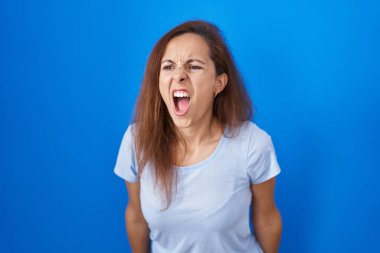 Brunette woman standing over blue background angry and mad screaming frustrated and furious, shouting with anger. rage and aggressive concept. 