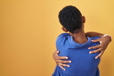 Sarı arka planda durup kendine sarılan Afrikalı Amerikalı kadın mutlu ve pozitiftir. Sevgi ve özgüven. 