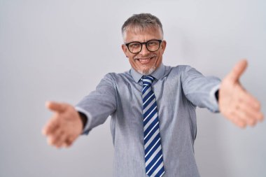 Hispanic business man with grey hair wearing glasses looking at the camera smiling with open arms for hug. cheerful expression embracing happiness. 