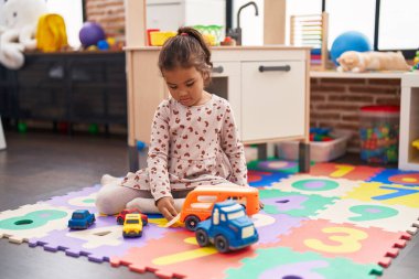 Anaokulunda yerde oturan sevimli İspanyol kız araba oyuncağıyla oynuyor.