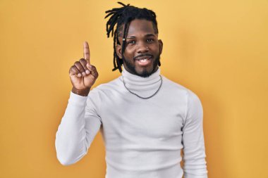 Sarı arka plan üzerine balıkçı yaka kazak giyen Afrikalı bir adam kendini beğenmiş ve mutlu gülümserken bir numaralı parmağıyla işaret ediyor.. 