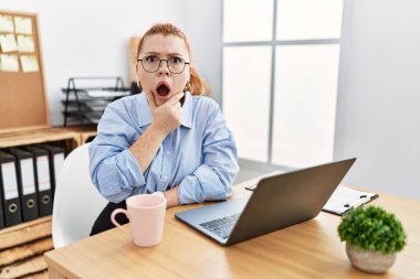 Young redhead woman working at the office using computer laptop looking fascinated with disbelief, surprise and amazed expression with hands on chin 