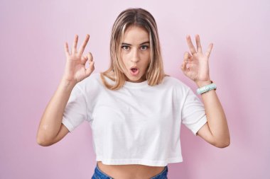 Young blonde woman standing over pink background looking surprised and shocked doing ok approval symbol with fingers. crazy expression 