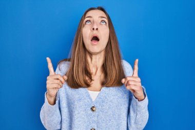 Mavi arka planda duran genç kadın şaşırmış ve şaşırmıştı. Parmaklarıyla işaret ediyor ve kollarını kaldırıyordu.. 