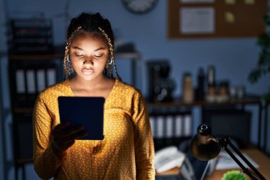African american woman with braids working at the office at night with tablet relaxed with serious expression on face. simple and natural looking at the camera. 