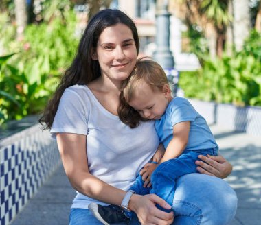 Anne ve oğlu parkta bankta otururken kendinden emin bir şekilde gülümsüyor.