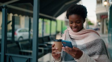Genç Afrikalı Amerikalı kadın akıllı telefon kullanıyor. Sokakta kahve içiyor.