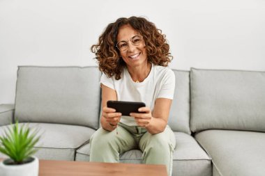 Orta yaşlı İspanyol kadın evde akıllı telefon kullanarak gülümsüyor.