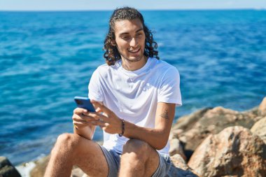 Young hispanic man using smartphone sitting on rock at seaside
