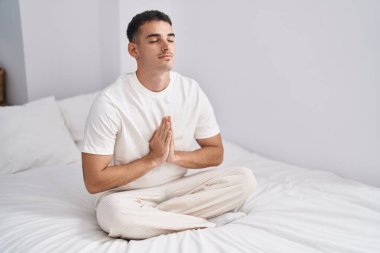 Young hispanic man doing yoga exercise sitting on bed at bedroom
