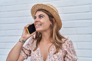 Young woman tourist wearing summer hat talking on smartphone over isolated white brick background
