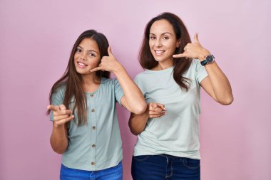 Young mother and daughter standing over pink background smiling doing talking on the telephone gesture and pointing to you. call me. 