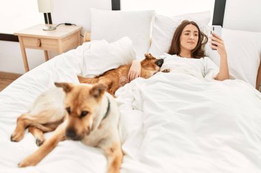 Genç İspanyol kadın köpeğe sarılıyor ve yatakta akıllı telefon kullanıyor.