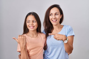 Young mother and daughter standing over white background smiling with happy face looking and pointing to the side with thumb up. 