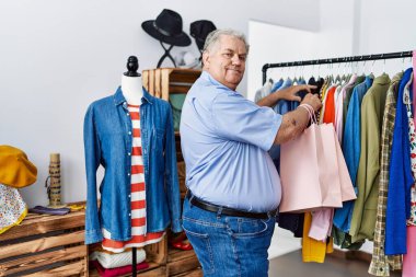Orta yaşlı, gri saçlı, gülümseyen, kendine güvenen bir adam giyim mağazasından alışveriş yapıyor.